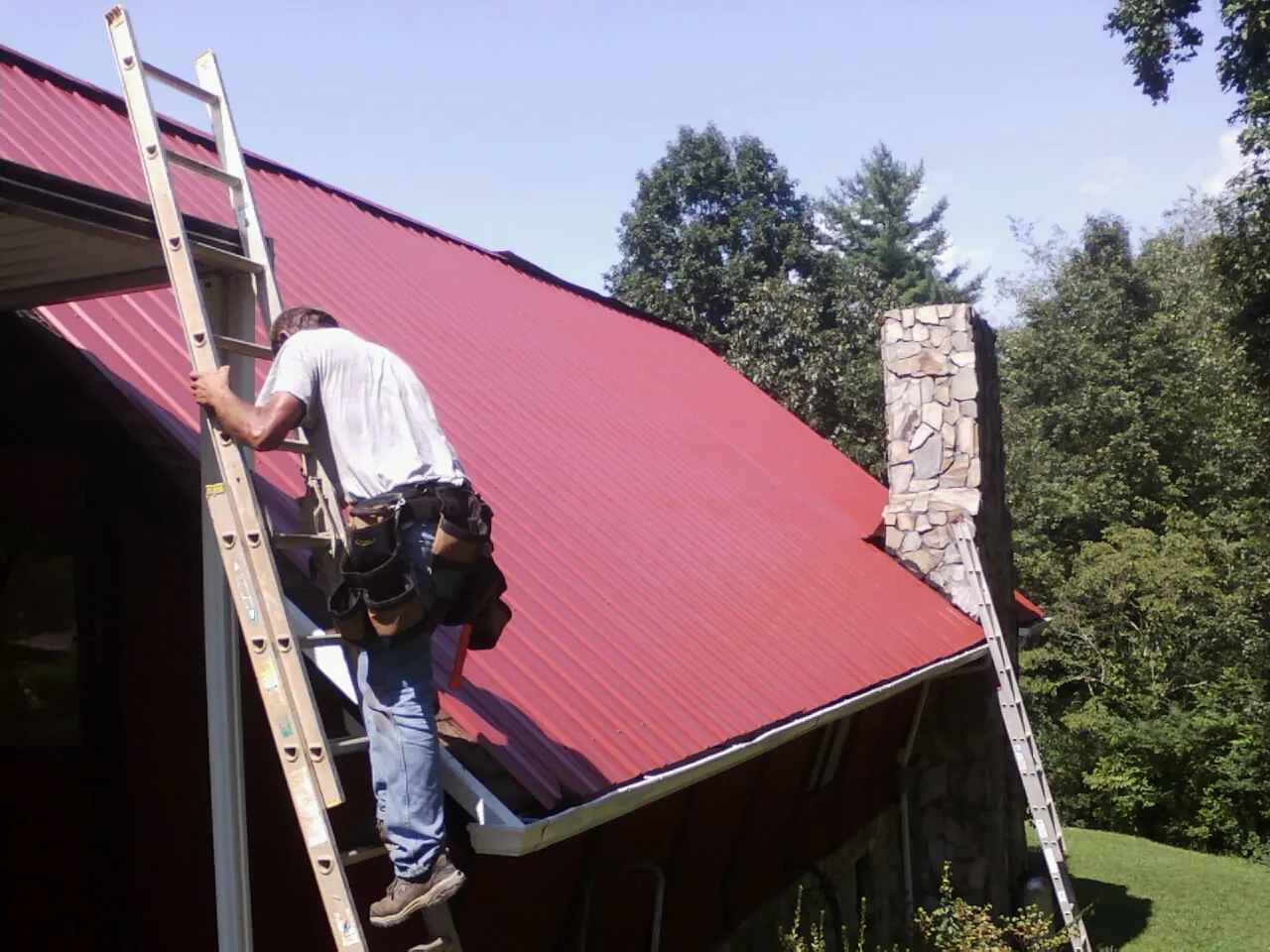 Roofer on a ladder beside a red metal roof and stone chimney — Soffit & Fascia Repair by Roofing Brothers in Eupora